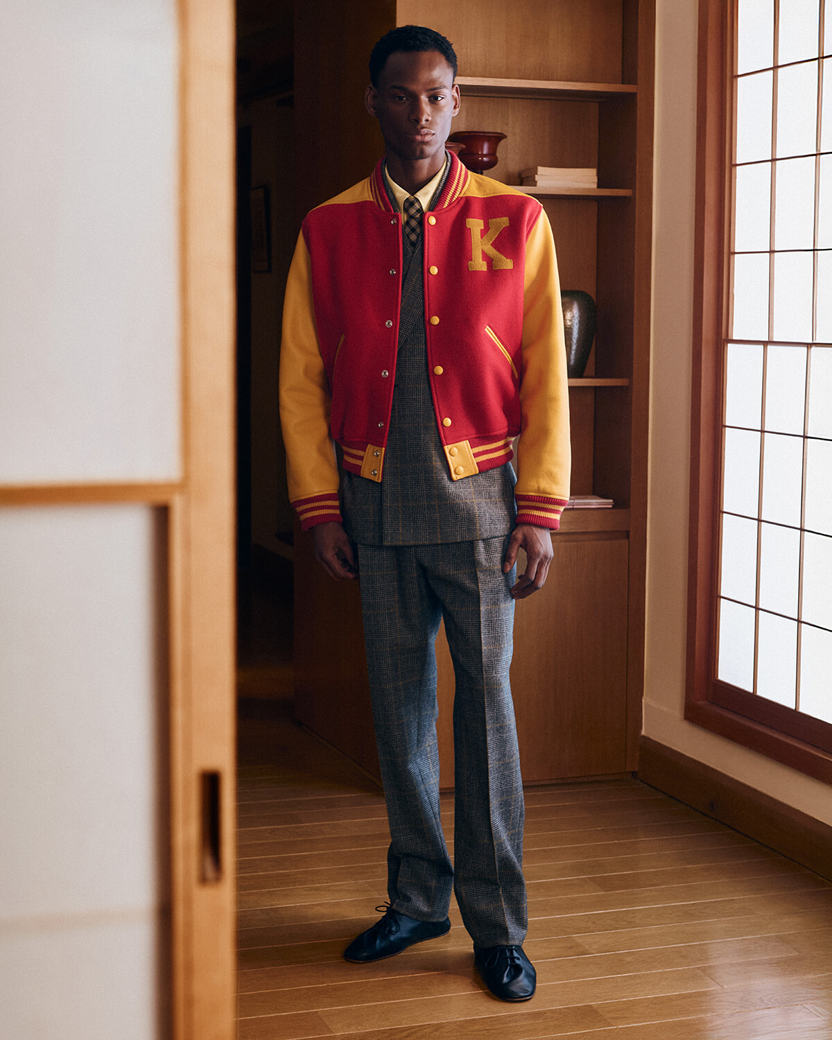 Young man standing in a doorway wearing a red and yellow varsity jacket over a gray plaid suit, indoors.