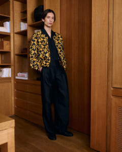 Model wearing a black outfit with a bold yellow geometric blazer, standing with hands in pockets against a wood-paneled wall and shelves.