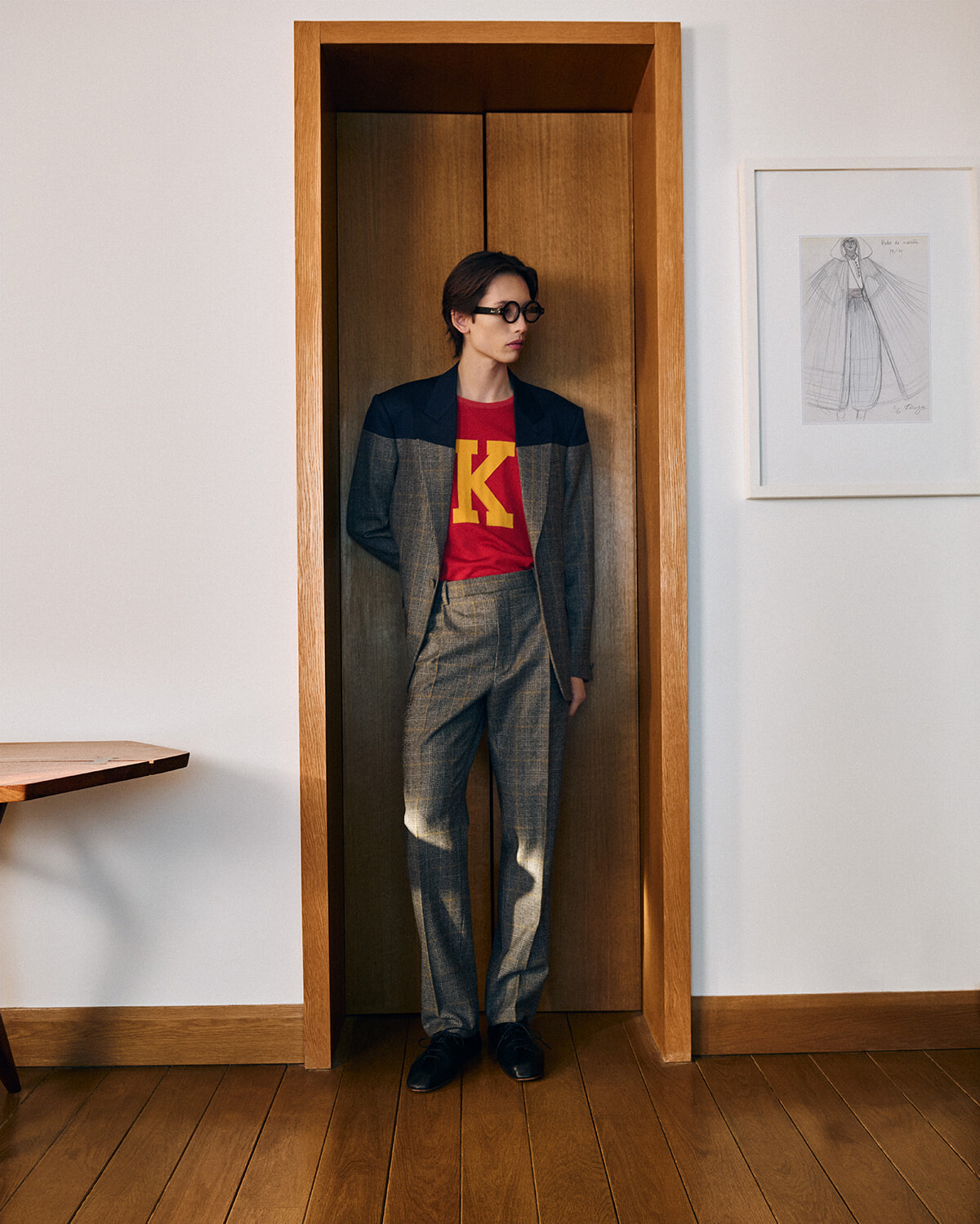 Person wearing a gray suit with a red shirt and sunglasses stands in an open wooden doorway, leaning slightly forward.