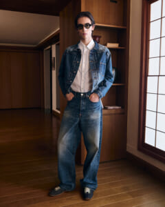 Person standing indoors in a denim-on-denim outfit: jacket over a light shirt, wide jeans, hands in pockets, wearing round sunglasses.