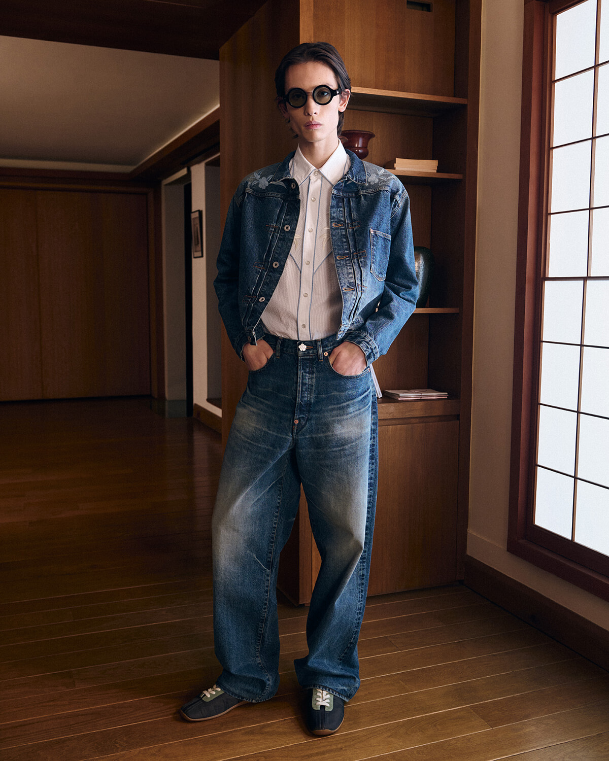 Person standing indoors in a denim-on-denim outfit: jacket over a light shirt, wide jeans, hands in pockets, wearing round sunglasses.