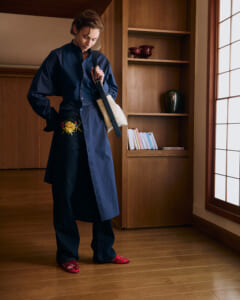 Person in a navy long coat with embroidered floral belt, standing indoors near a wooden bookshelf, carrying a beige shoulder bag and wearing red floral slippers.
