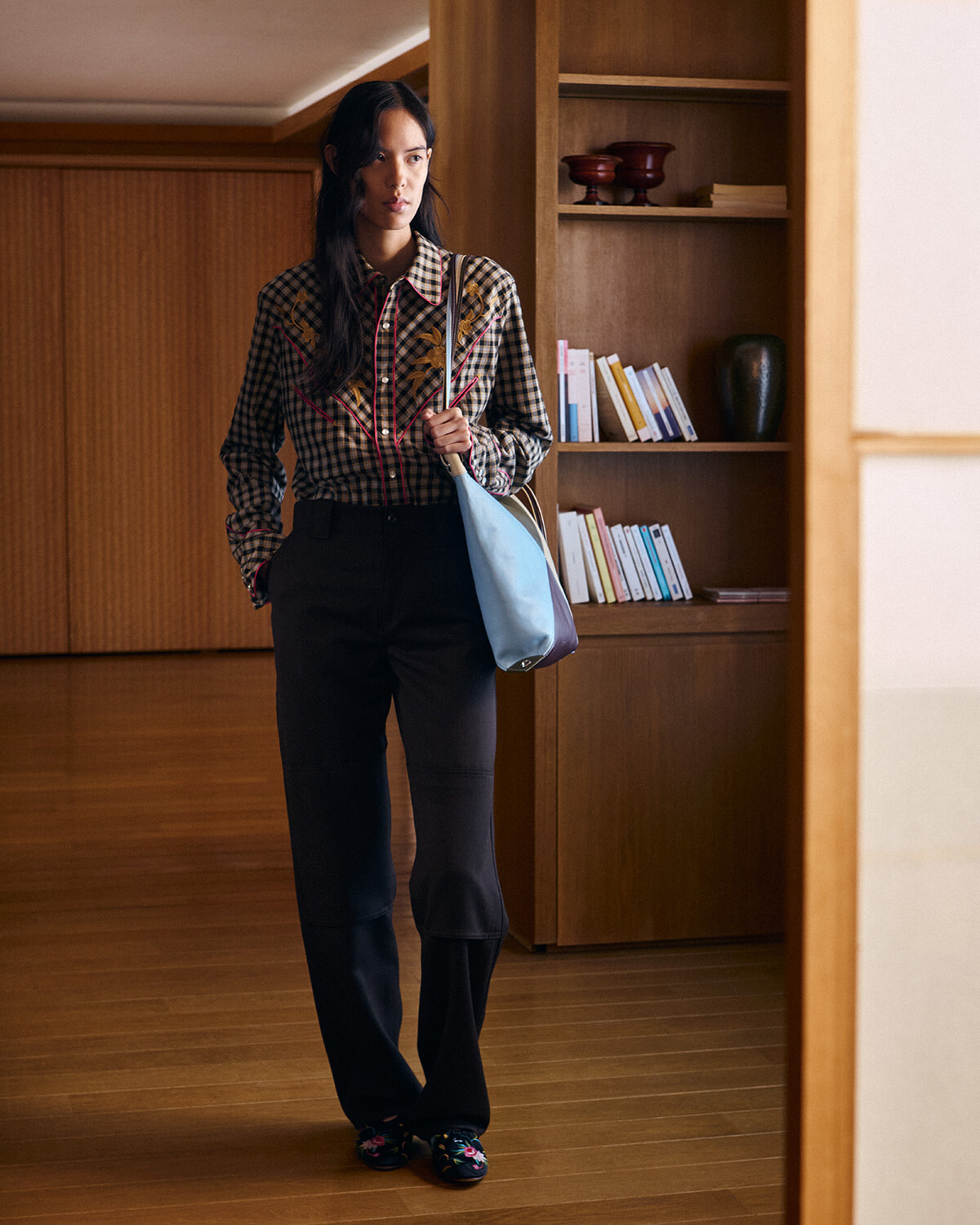 Woman in a plaid embroidered shirt and black wide‑leg pants stands in a wood-paneled room, carrying a light blue bag over her shoulder.