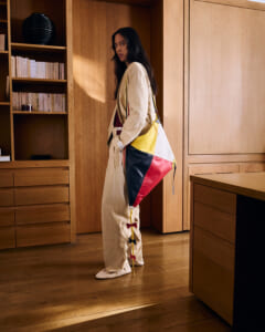 Woman in a beige jumpsuit poses in a wood-furnished room, carrying a tri-color patchwork bag (yellow, white, red, navy) and looking at the camera.