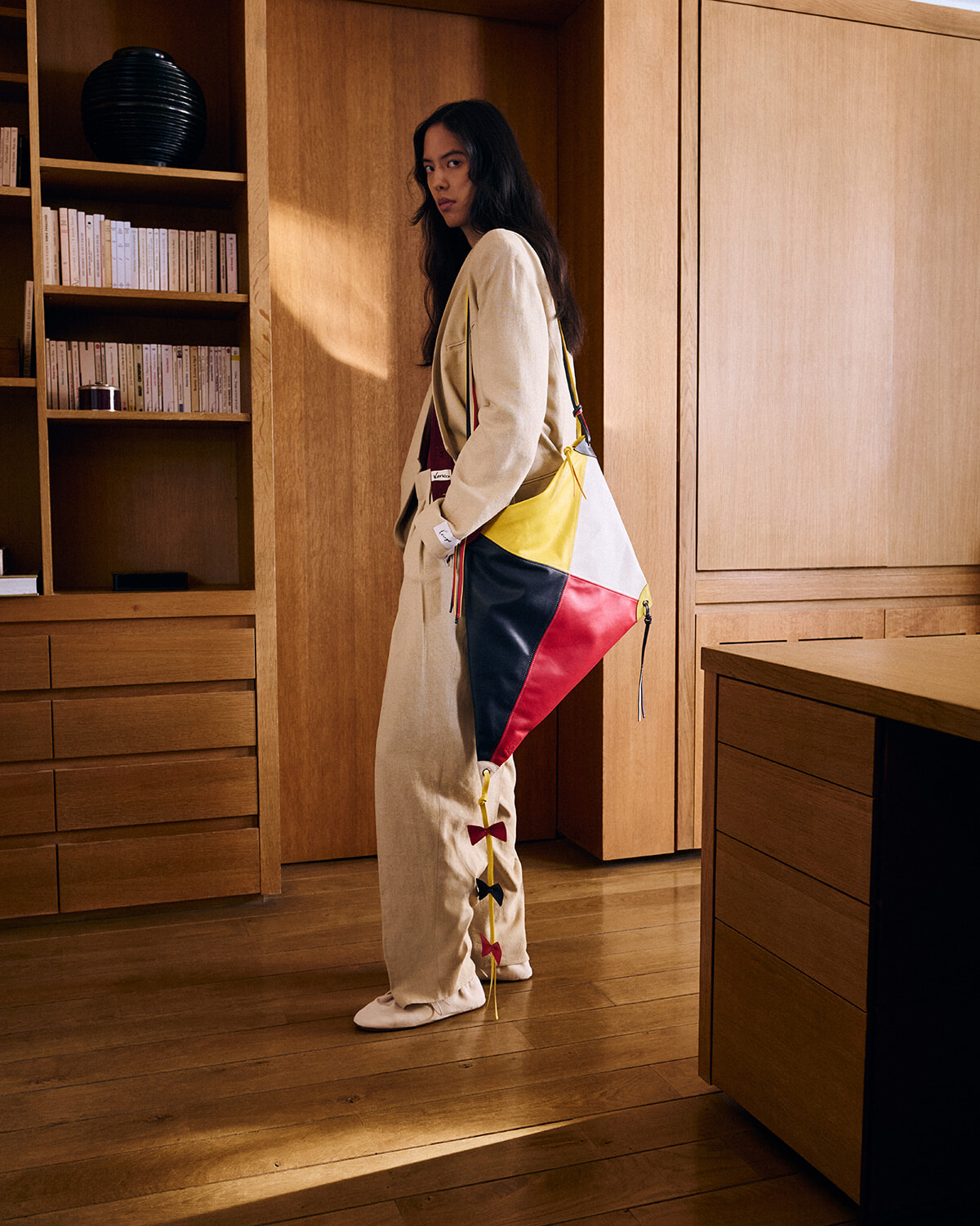 Woman in a beige jumpsuit poses in a wood-furnished room, carrying a tri-color patchwork bag (yellow, white, red, navy) and looking at the camera.