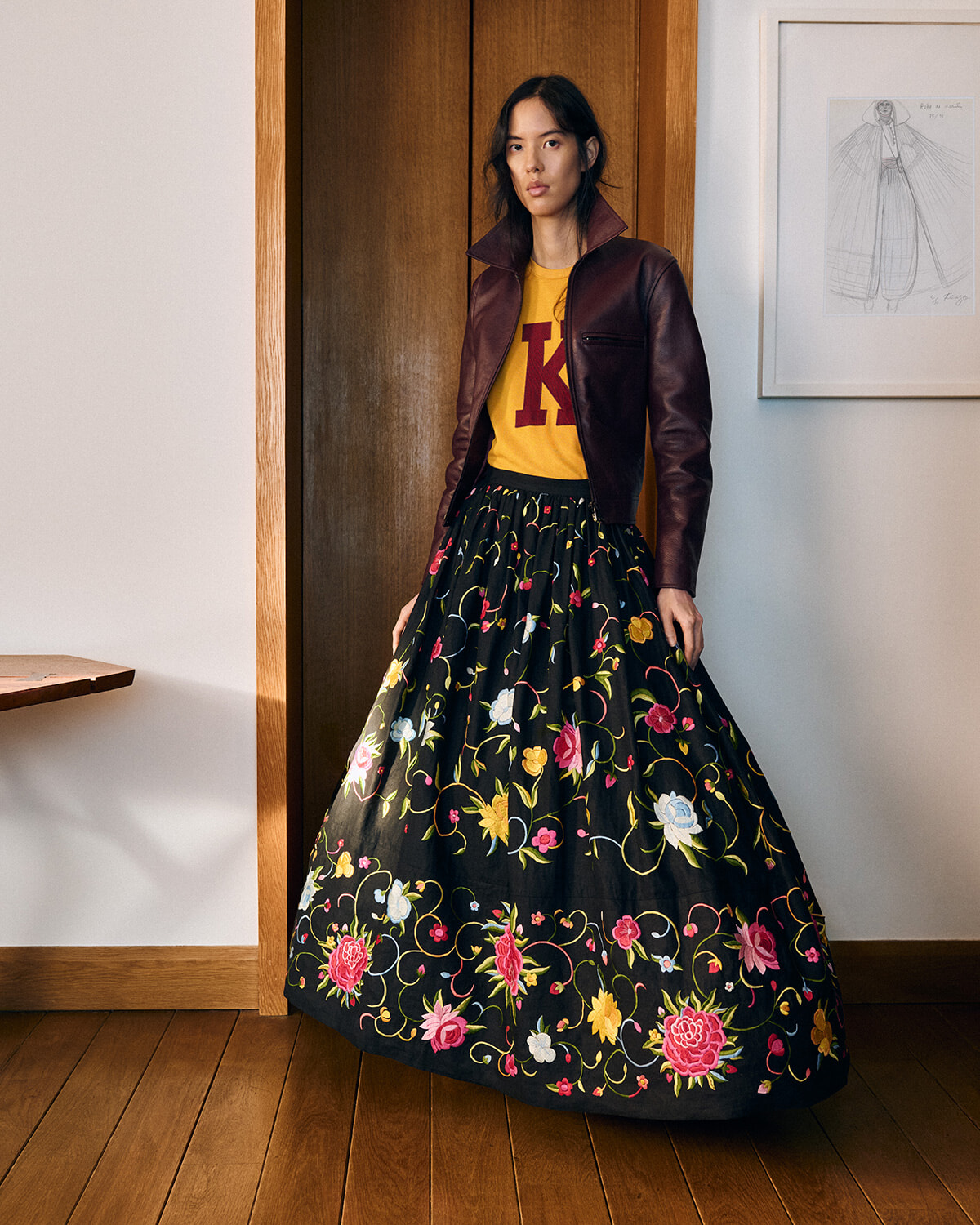 Full-length portrait of a woman indoors wearing a burgundy leather jacket, a mustard yellow top, and a long black floral skirt.