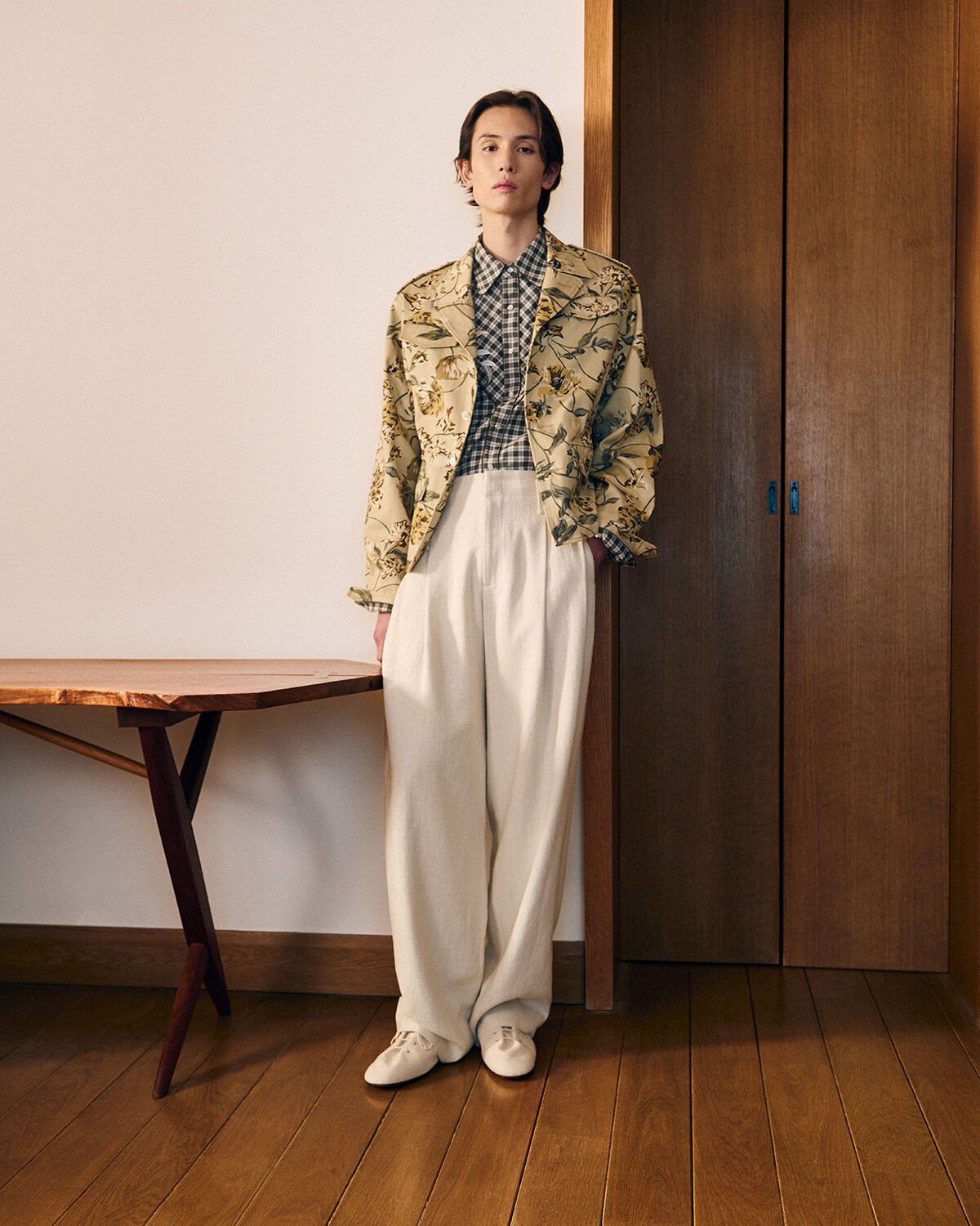 Person standing against a wall in a beige floral blazer, checkered shirt, and wide cream trousers with white shoes.