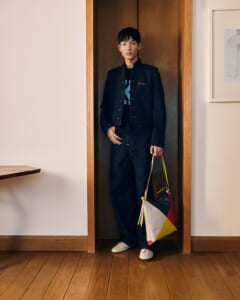 Young man standing in a doorway wearing a dark denim jacket and pants, holding a colorful geometric handbag.