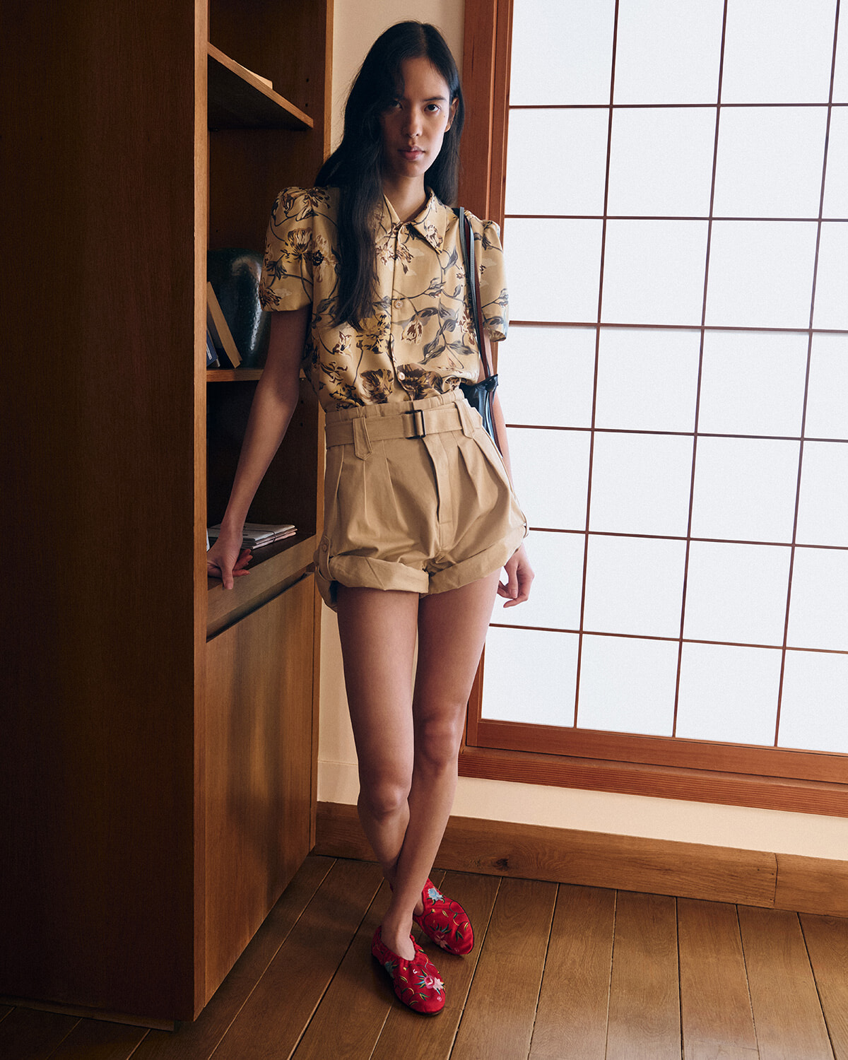 Woman standing indoors by a wooden bookshelf and shoji screen, wearing a beige floral blouse and high-waisted khaki shorts with red floral slippers.