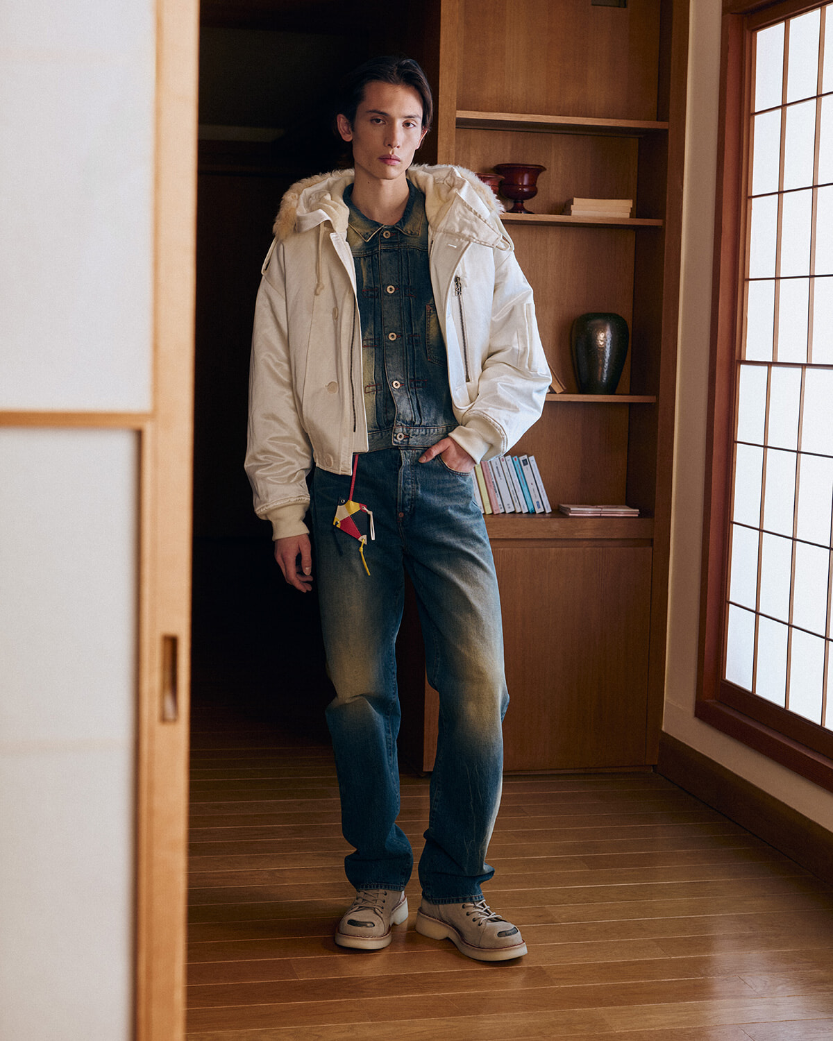 Person standing in a doorway wearing a white fur-edged jacket over a denim jacket and blue jeans, hands in pockets.