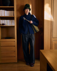 Person in a navy coat and green checkered hat stands by a wooden cabinet, carrying a beige messenger bag.