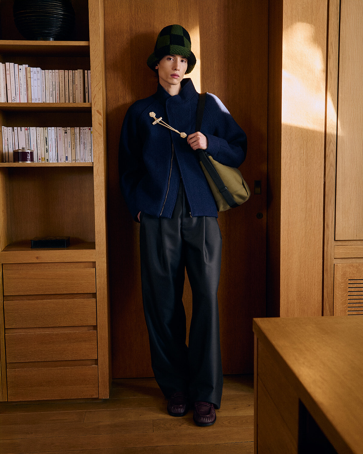 Person in a navy coat and green checkered hat stands by a wooden cabinet, carrying a beige messenger bag.