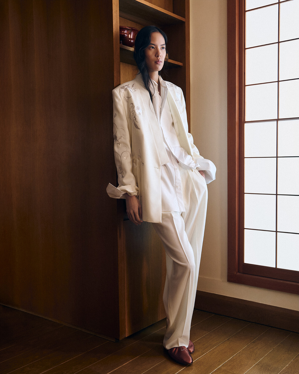 Model in an all-white suit with embroidered blazer leaning against a wooden cabinet by a sunlit windowed wall.