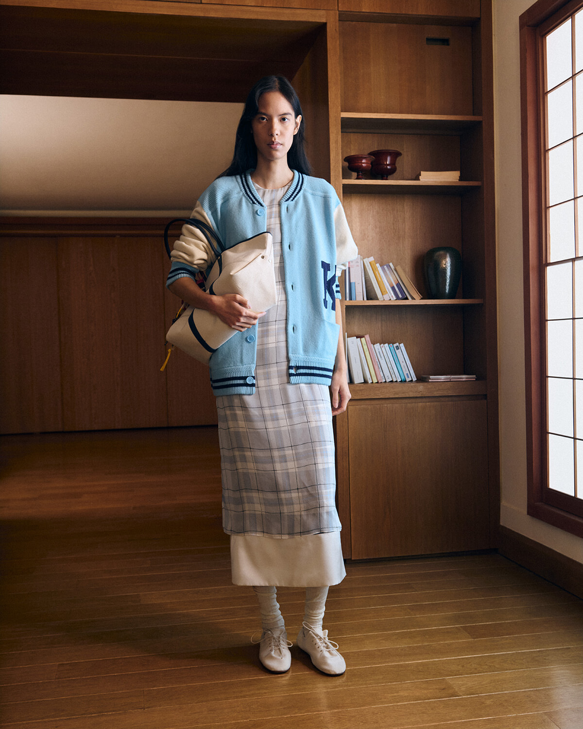 Young woman standing in a warm wooden interior wearing a light blue varsity jacket over a plaid dress, holding a beige bag.