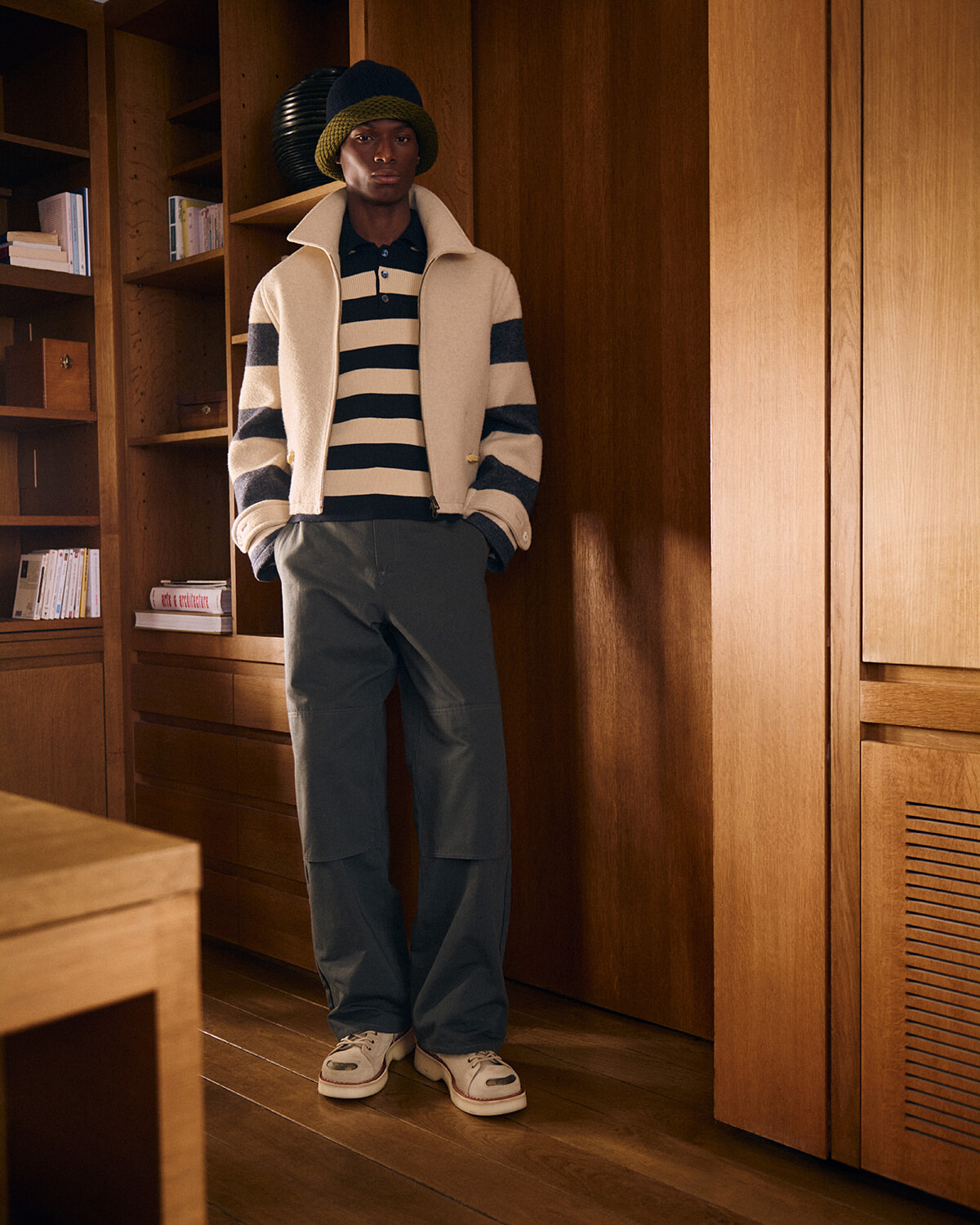 Man modeling a retro-inspired outfit: cream zip jacket with dark stripes, navy striped polo, green beanie, and dark cargo pants indoors among wooden shelves and cabinetry.