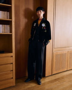 Young man in a black velvet jacket with floral embroidery stands with hands in pockets in a wooden room.