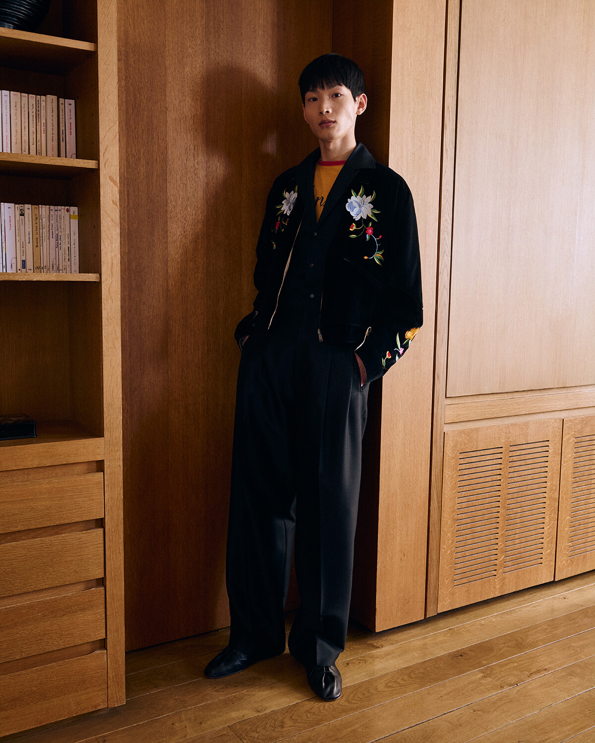 Young man in a black velvet jacket with floral embroidery stands with hands in pockets in a wooden room.