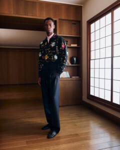 Man standing in a wood-paneled room wearing a black floral sweater over a pink shirt and black trousers.