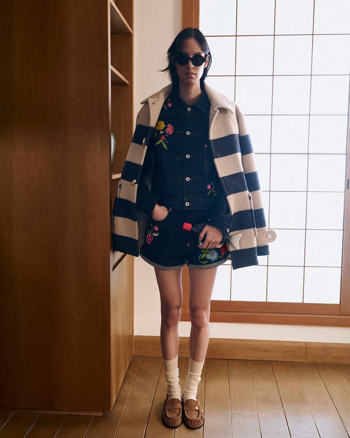 Person indoors wearing a floral-embroidered denim outfit with a cream-and-navy striped coat draped over shoulders, sunglasses, and brown loafers with white socks.