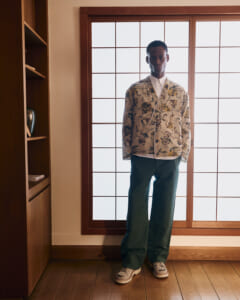 Man standing indoors by a wooden-framed window, wearing a cream floral print jacket, white shirt, green pants, and sneakers.