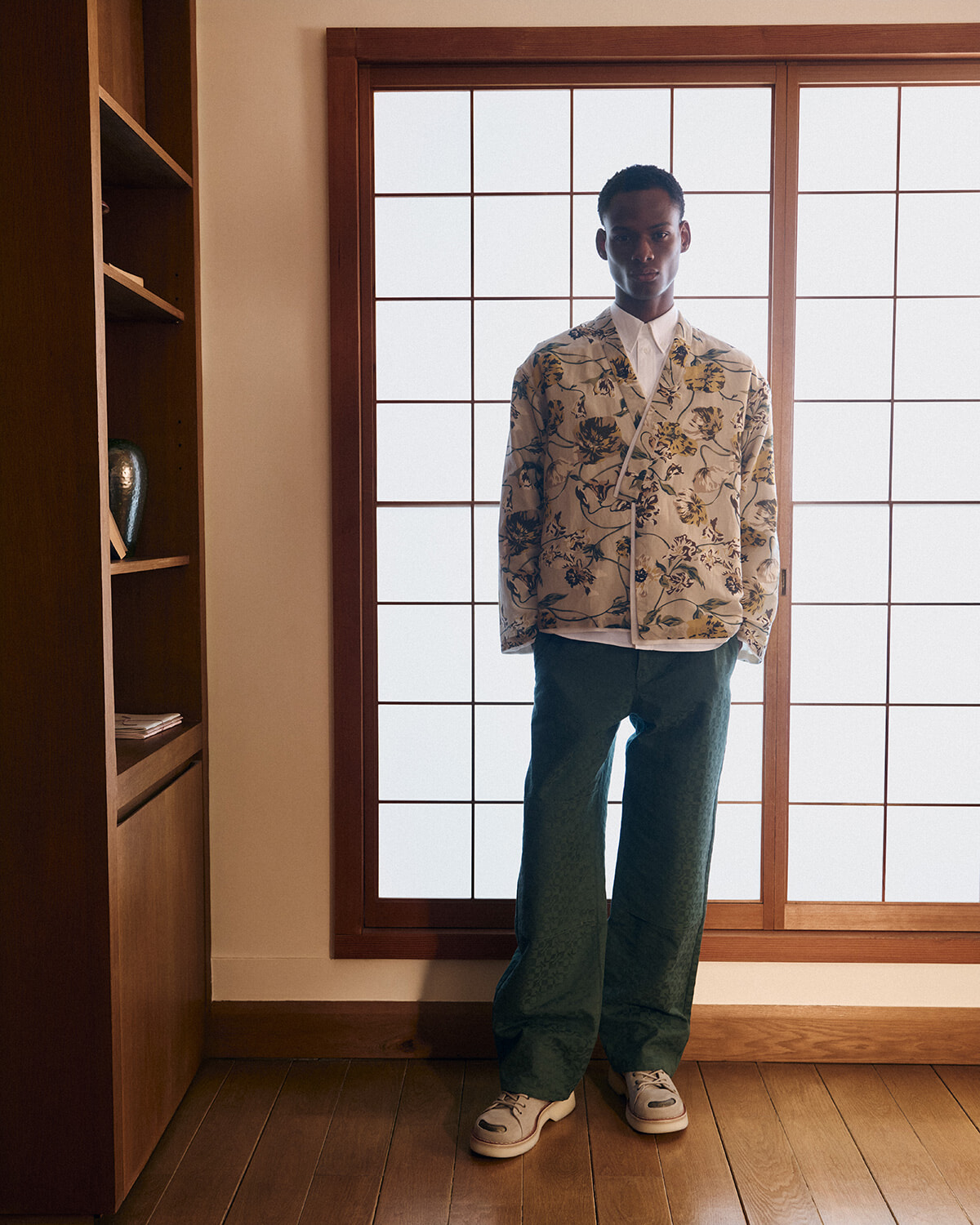 Man standing indoors by a wooden-framed window, wearing a cream floral print jacket, white shirt, green pants, and sneakers.