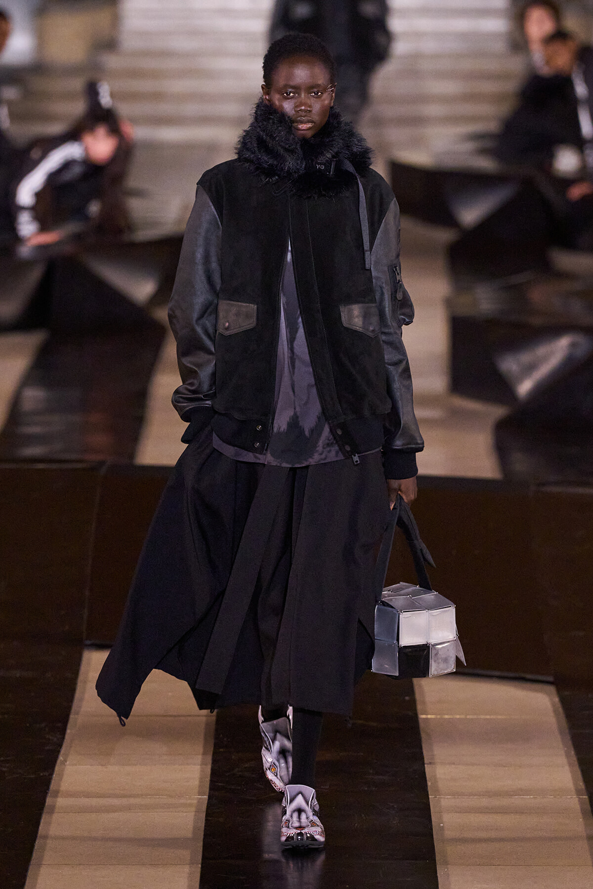 Model walks a runway in an oversized black coat with a fur collar, layered over a dark skirt, carrying a metallic cube handbag.
