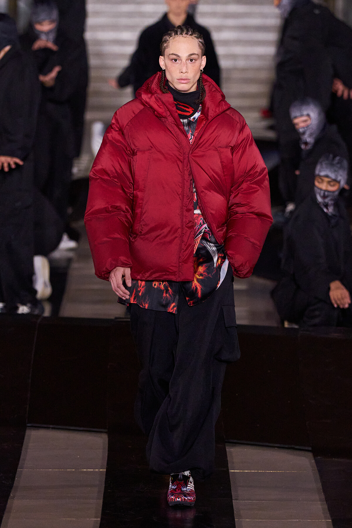 Model walking the runway in an oversized red puffer jacket over a graphic top and black cargo pants, with cornrow braids, on a glossy catwalk.