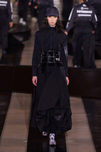 Model walks a dark, avant-garde runway in a black zip-top, layered skirt, glossy patent belt with Y-3 buckle, and a black cap.
