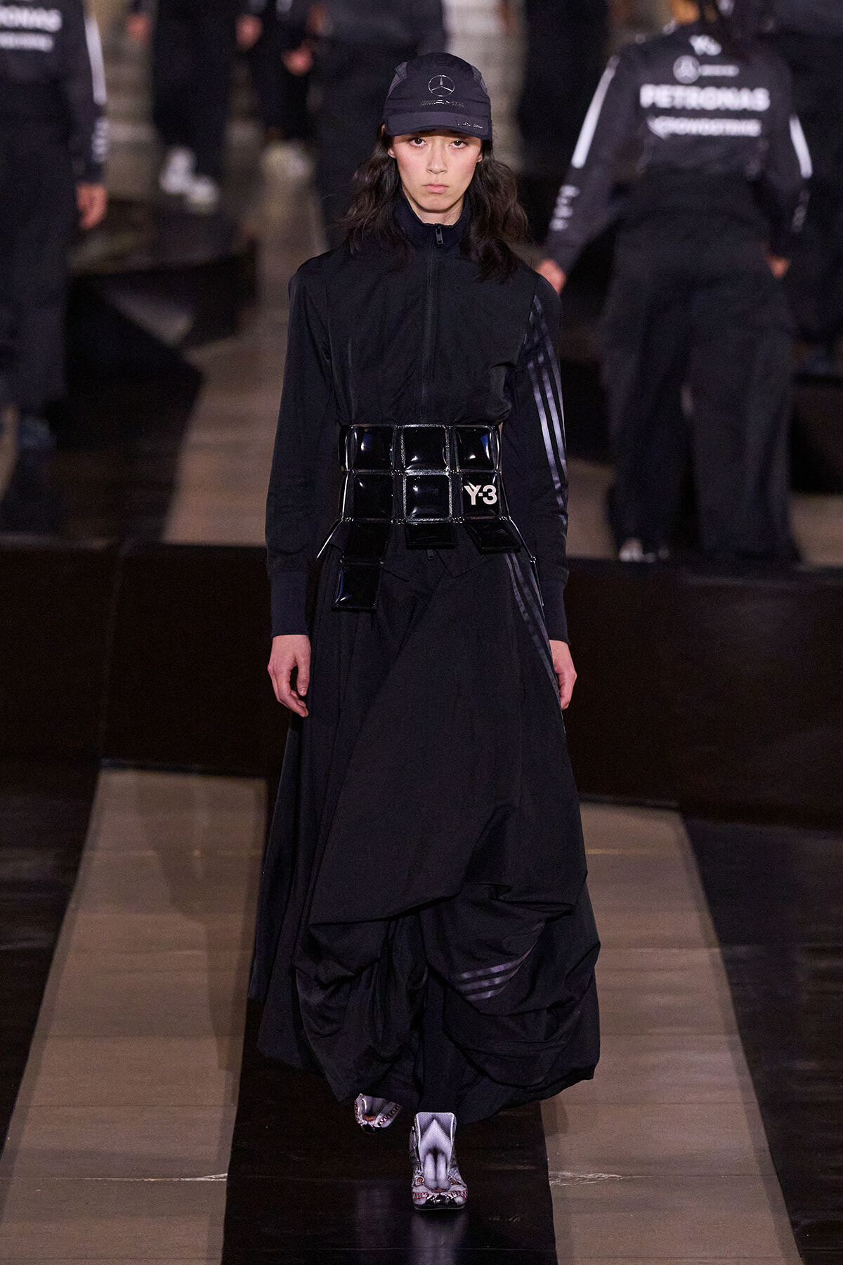 Model walks a dark, avant-garde runway in a black zip-top, layered skirt, glossy patent belt with Y-3 buckle, and a black cap.