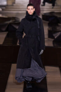 Female model walking a fashion runway in a black belted trench with a fur collar and a voluminous gray skirt on the catwalk.