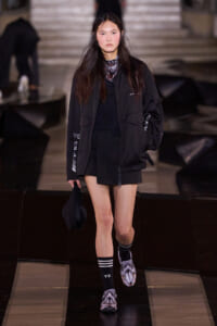 Model walks a dark runway in an all-black streetwear look: oversized jacket, black shorts, striped socks, and wolf-face slippers, holding a black bag.