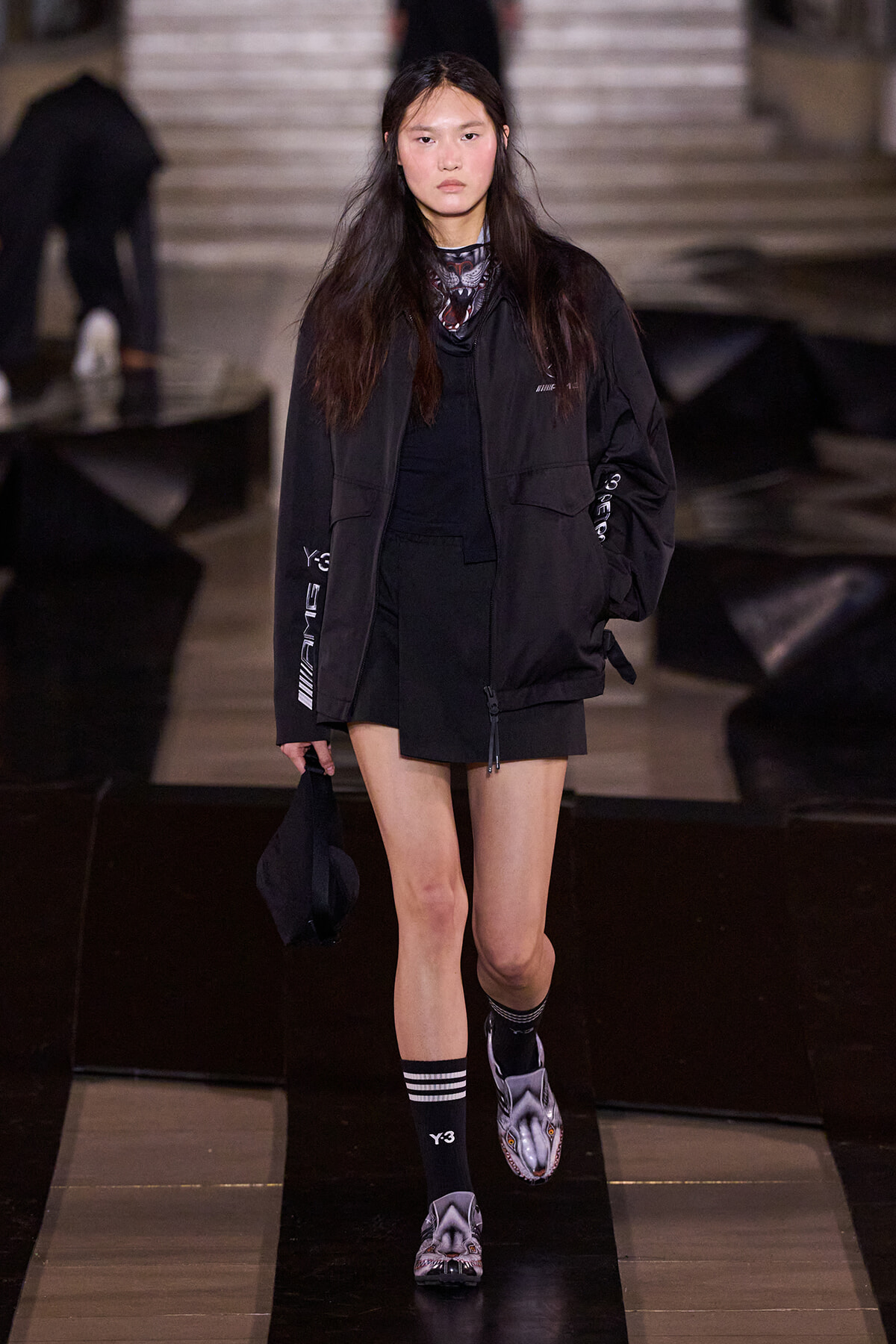 Model walks a dark runway in an all-black streetwear look: oversized jacket, black shorts, striped socks, and wolf-face slippers, holding a black bag.