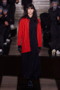 Runway model walking the catwalk in a bright red textured cardigan over a long black shirt and wide black pants, wearing black gloves and sneakers, with masked audience in the background.