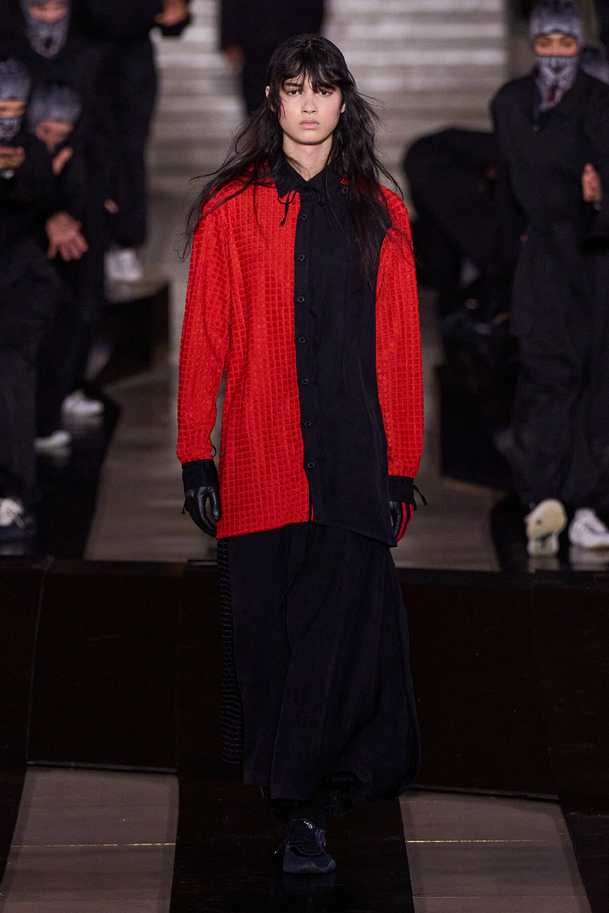 Runway model walking the catwalk in a bright red textured cardigan over a long black shirt and wide black pants, wearing black gloves and sneakers, with masked audience in the background.