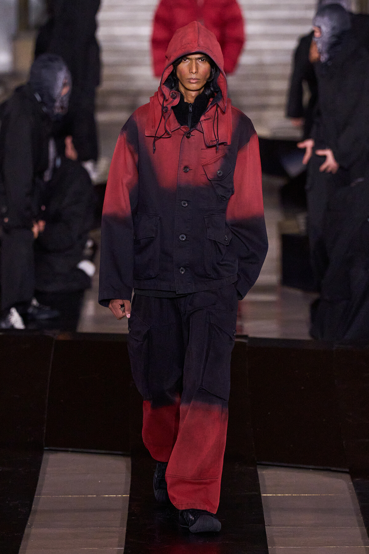 Model wearing a red-to-black gradient hooded jacket with matching cargo pants on a fashion runway., Female model in a color-block red and black parka with oversized pockets walking the catwalk., Runway look featuring a red-to-black gradient hooded jacket and loose cargo pants., Fashion show model in an urban-inspired red-dipped parka and black pants, walking the runway., Gradient red and black outerwear with hood, paired with wide cargo trousers, on a fashion show.