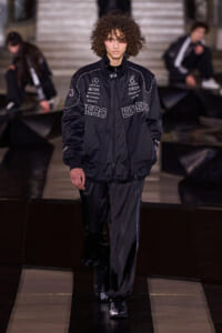 Male model with curly hair walking a fashion runway in a black logo-covered tracksuit, center stage.