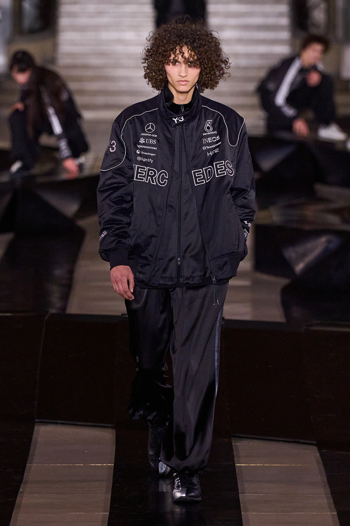 Male model with curly hair walking a fashion runway in a black logo-covered tracksuit, center stage.