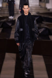 Male model walks the runway in an all-black outfit: suede-like jacket with leather sleeves and shiny leather pants.