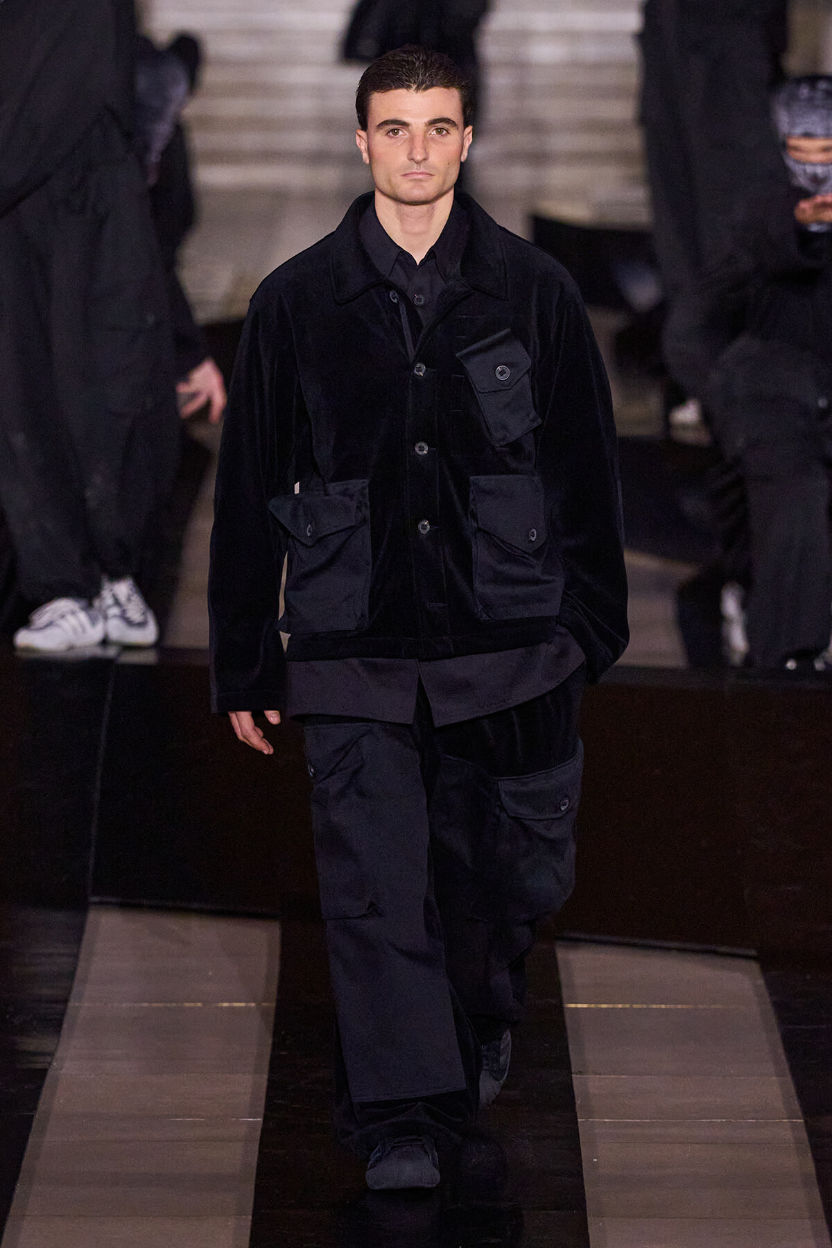 Male model walking a runway in a black velvet utility jacket with multiple pockets and large pockets on the chest and hips.