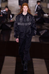 Model with red hair walking a fashion runway in a black Mercedes‑AMG jacket with logos, black pants, and boots.