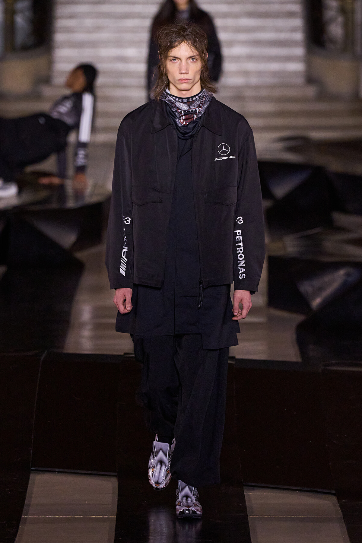 Male model on a runway wearing an oversized black jacket with AMG and Petronas branding and layered black pants, silver-patterned boots.