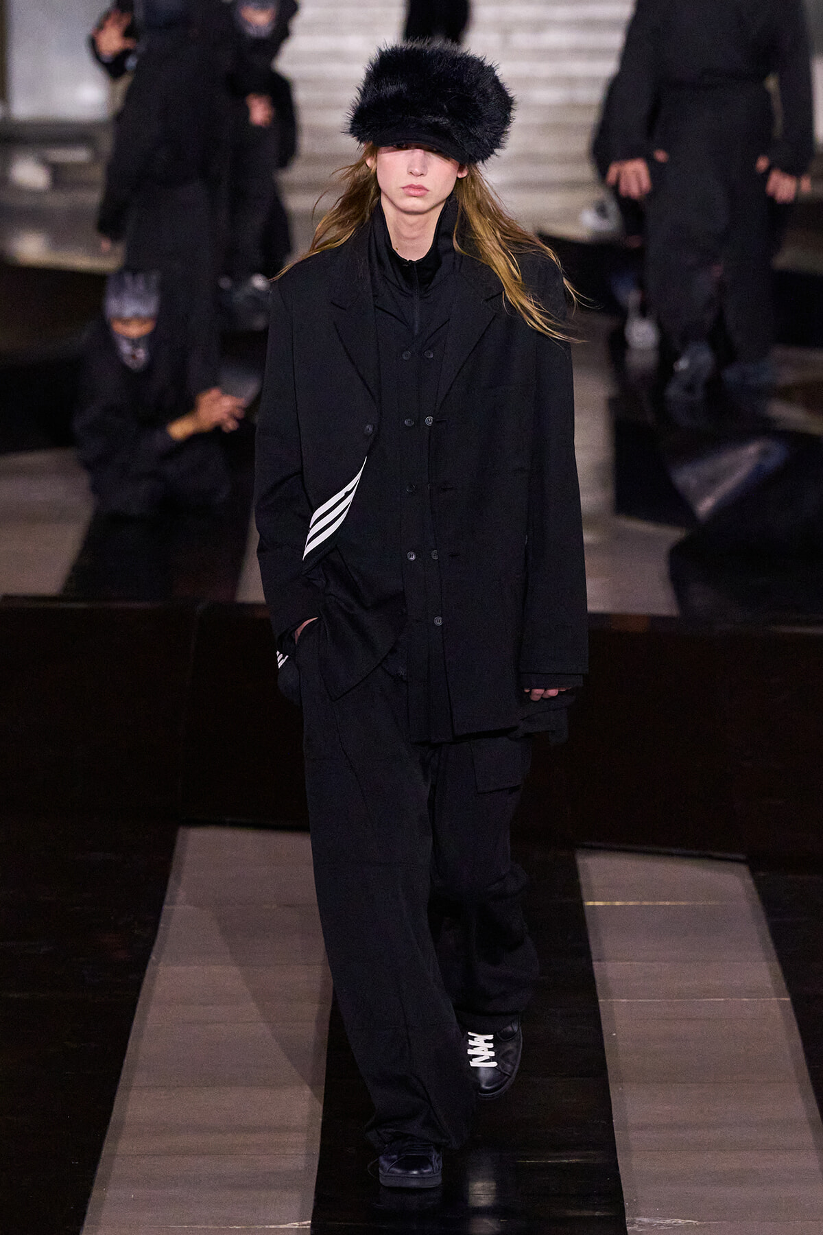 Runway model in an all-black layered outfit with a large black fur hat, walking the catwalk with hands in pockets.