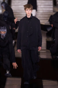 Male model walks a dark, oversized black ensemble on a runway, with masked assistants in the background.