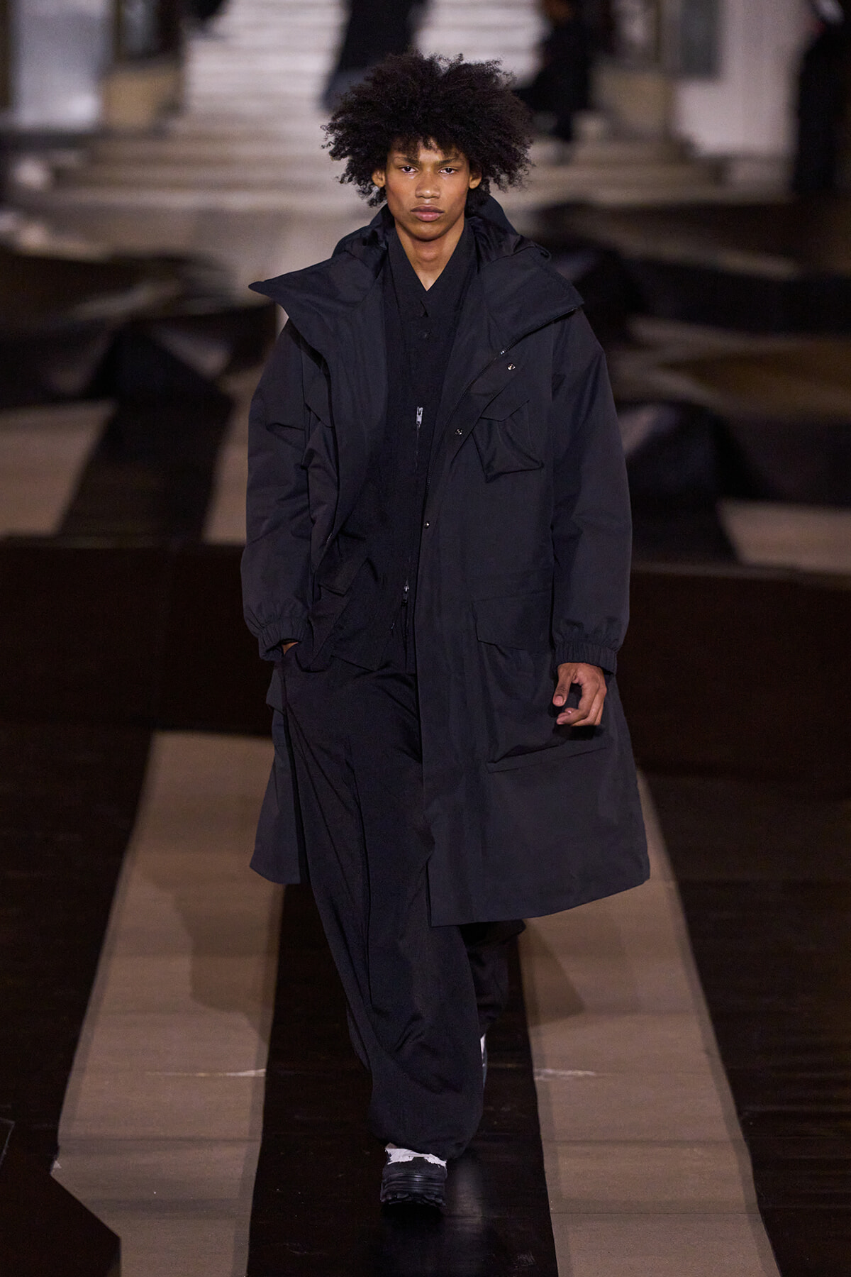 Male model wearing an oversized black parka with multiple pockets walks the fashion runway at nightfall settings.
