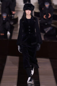 Model walking a runway in an all-black velvet suit with a fur hat, black gloves, and black-and-white sneakers.