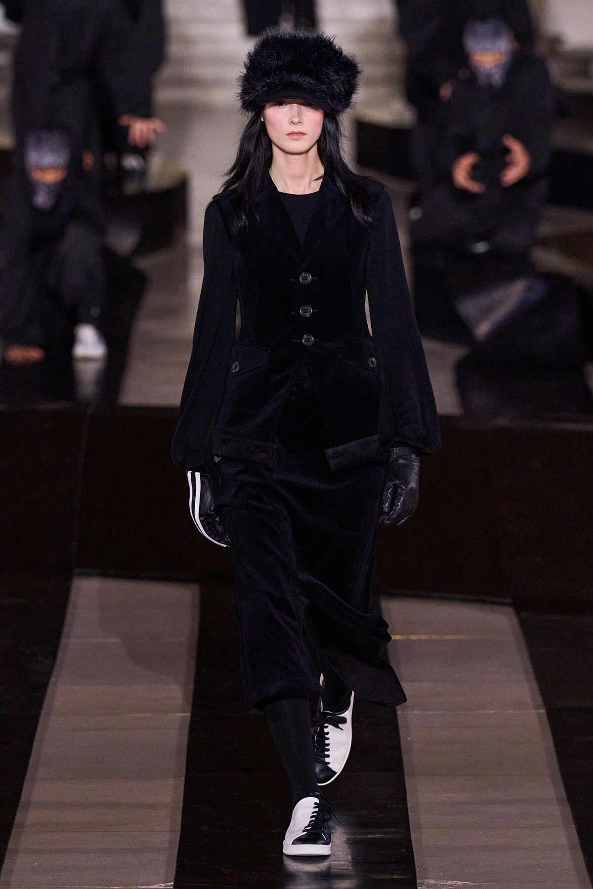 Model walking a runway in an all-black velvet suit with a fur hat, black gloves, and black-and-white sneakers.