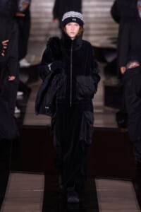 Runway model in an all-black velour outfit with oversized pockets, carrying a fur stole and wearing a black Y-3 knit beanie.
