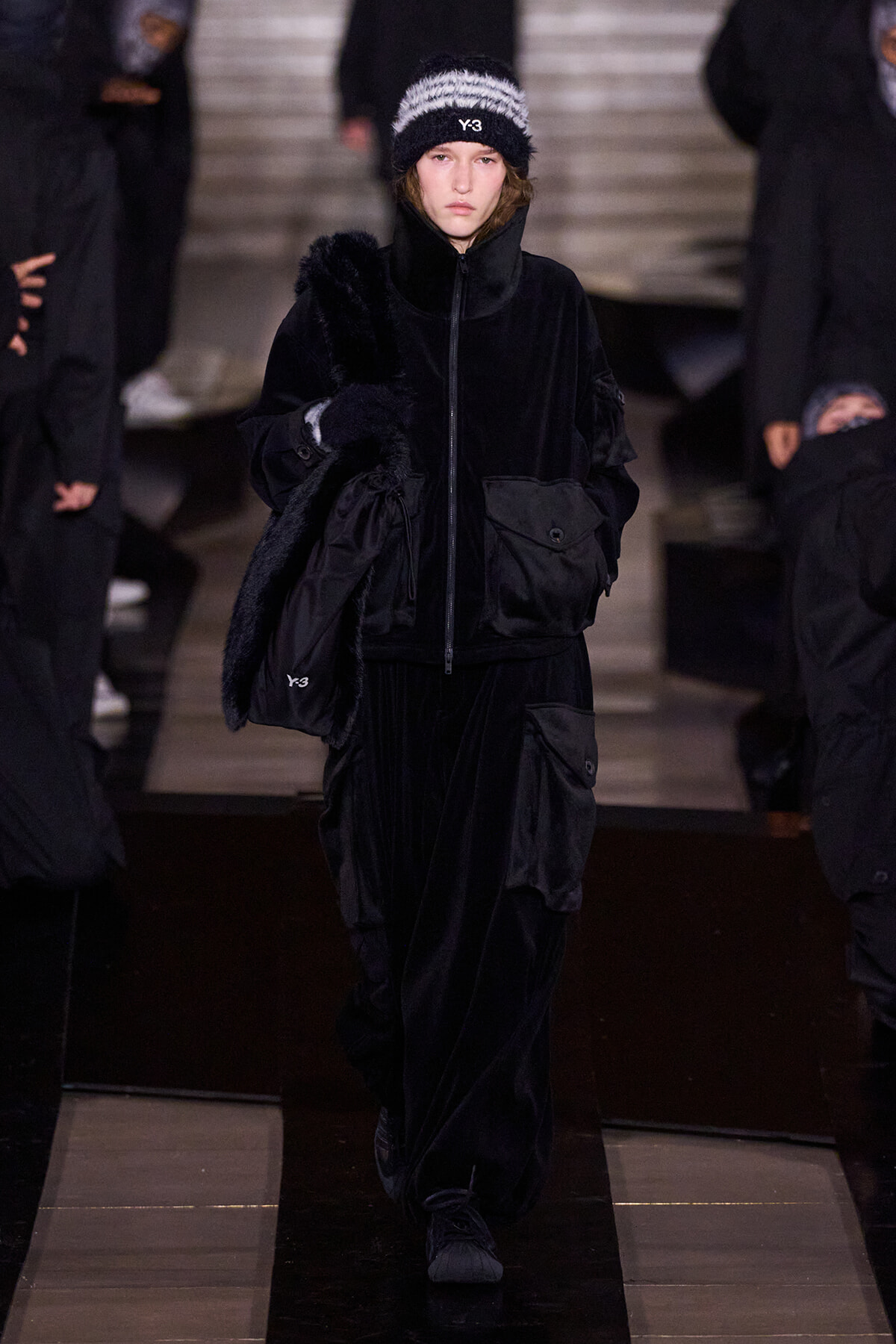 Runway model in an all-black velour outfit with oversized pockets, carrying a fur stole and wearing a black Y-3 knit beanie.