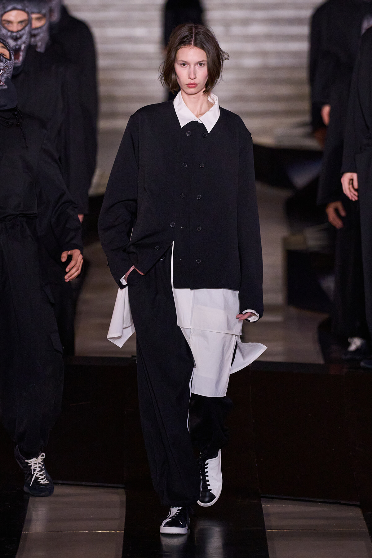 Model walking a runway in an oversized black double-breasted jacket over a white shirt with layered white and black trousers and black-and-white sneakers; backstage-like dark backdrop.