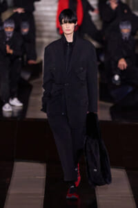 Model walks a runway in an oversized black utility jacket with multiple pockets, black pants, red sneakers, and a black fur-lined bag; dark hair clipped at the bangs with a focused expression.